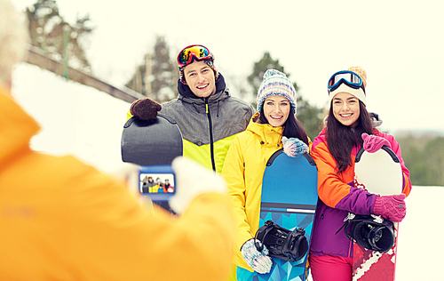 winter sport, technology, leisure, friendship and people concept - happy friends with snowboards and smartphone taking picture outdoors