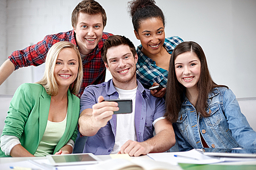 education, people, friendship, technology and learning concept - group of happy international high school students or classmates with smartphone taking selfie in classroom