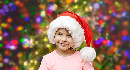 holidays, christmas, childhood and people concept - smiling little girl in santa hat sitting on couch over glitter or lights background