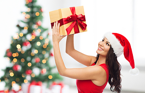 people, holidays, christmas and celebration concept - beautiful  woman in red dress and santa hat with gift box over room with christmas tree and gifts background