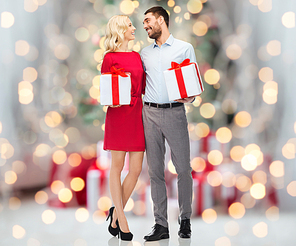 people, christmas, birthday, couple and holidays concept - happy young man and woman with gift boxes over christmas tree lights background