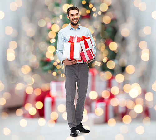 people, christmas, birthday and holidays concept - happy young man holding gift boxes over christmas tree lights background