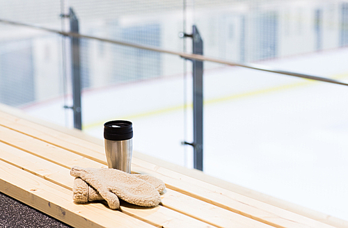 hot drink, winter and leisure concept - close up of thermos cup and sheepskin mittens on bench at ice rink arena