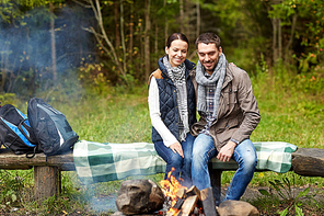 camping, travel, tourism, hike and people concept - happy couple sitting on bench and warming near campfire at camp in woods