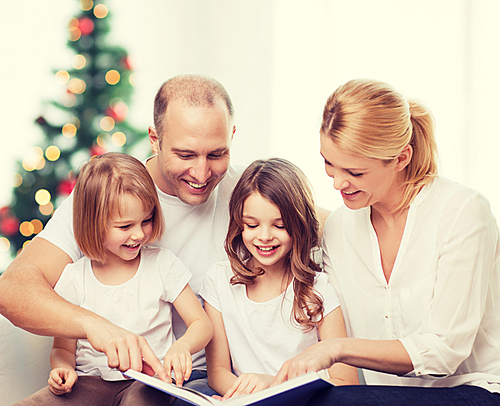 family, childhood, holidays and people - smiling mother, father and little girls reading book over living room and christmas tree background