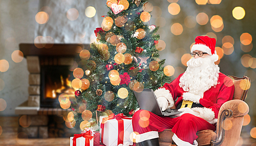 technology, holidays and people concept - man in costume of santa claus with laptop computer, gifts and christmas tree sitting in armchair over home fireplace and lights background