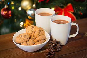 holidays, winter, food and drinks concept - close up of oatmeal cookies, cups with hot chocolate or cocoa drink and pinecones on wooden table over christmas tree background