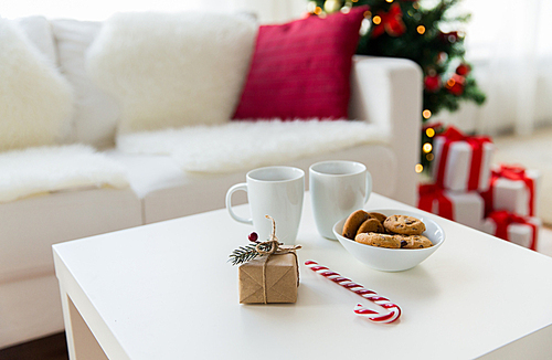 christmas, holidays and  winter concept - close up of gift, oat cookies, sugar cane candy and cups on table at home