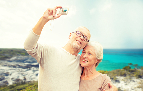 age, tourism, travel, technology and people concept - senior couple with camera taking selfie on street over beach background