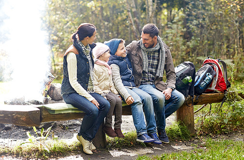 camping, travel, tourism, hike and people concept - happy family sitting on bench and talking at camp in woods