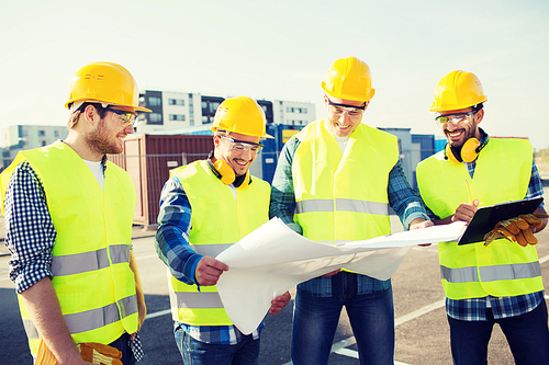 business, building, teamwork and people concept - group of smiling builders in hardhats with clipboard and blueprint outdoors