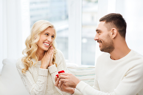 love, couple, relationship, proposal and holidays concept - happy man giving engagement ring in little red gift box to woman at home