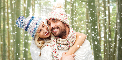 winter, fashion, couple, christmas and people concept - smiling man and woman in hats and scarf hugging over snowflakes and forest background