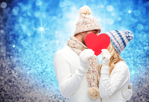 love, valentines day, couple, christmas and people concept - smiling man and woman in winter hats and scarf hiding behind red paper heart shape over blue glitter and holidays lights background