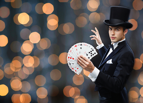 magic, gambling, casino, people and show concept - magician in top hat showing trick with playing cards over nigh lights background
