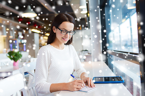 leisure, business, people, technology and lifestyle concept - smiling young woman in eyeglasses with tablet pc computer and notebook taking notes at cafe with snow effect