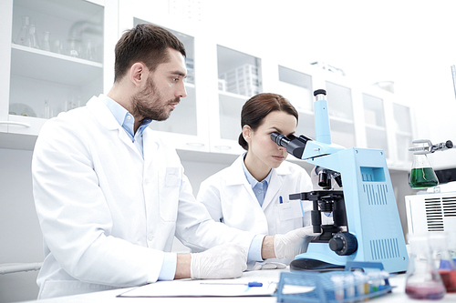 science, chemistry, technology, biology and people concept - young scientists with microscope making test or research in clinical laboratory and taking notes