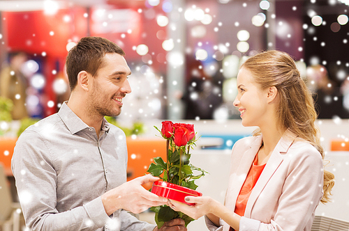love, romance, valentines day, couple and people concept - happy young man with red flowers giving present to smiling woman at cafe in mall with snow effect