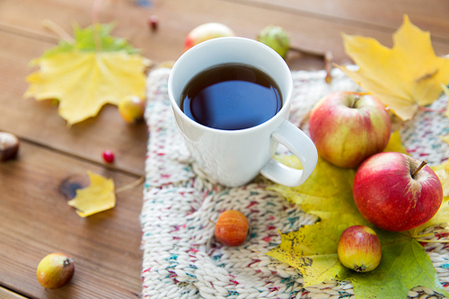 season, drink and morning concept - close up of tea cup on wooden table with autumn leaves and scarf