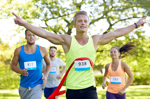 fitness, sport, victory, success and healthy lifestyle concept - happy man winning race and coming first to finish red ribbon over group of sportsmen running marathon with badge numbers outdoors