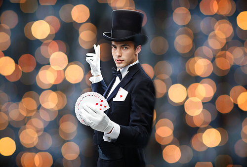 magic, performance, gambling, casino, people and show concept - magician in top hat showing trick with playing cards over nigh lights background