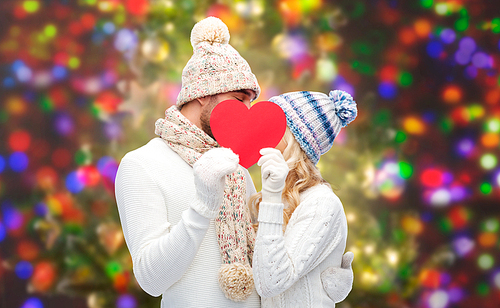love, valentines day, couple, christmas and people concept - smiling man and woman in winter hats and scarf hiding behind red paper heart shape over holidays lights background