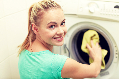 people, housework, laundry and housekeeping concept - happy woman putting laundry into washing machine at home