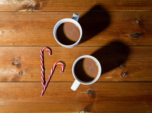 holidays, christmas, winter, food and drinks concept - close up of candy canes and cups with hot chocolate or cocoa drinks on wooden table