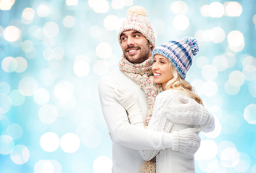 winter, fashion, couple, christmas and people concept - smiling man and woman in hats and scarf hugging over blue holidays lights background