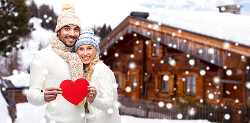 love, valentines day, couple, christmas and people concept - smiling man and woman in winter hats and scarf holding red paper heart shape over wooden country house and snow background