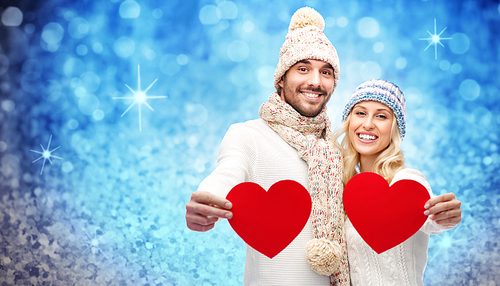 love, valentines day, couple, christmas and people concept - smiling man and woman in winter hats and scarf holding red paper heart shapes over blue glitter or lights background