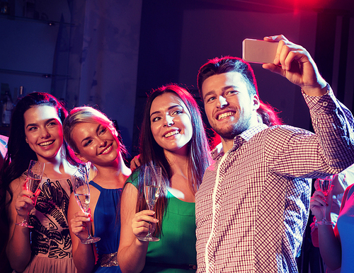 party, holidays, technology, nightlife and people concept - smiling friends with glasses of champagne and smartphone taking selfie in club