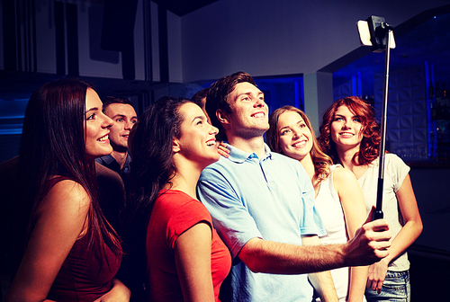 party, technology, nightlife and people concept - smiling friends with smartphone and monopod taking selfie in club