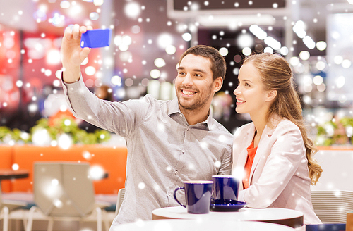 sale, shopping, consumerism, technology and people concept - happy young couple with smartphone taking selfie and drinking coffee or tea at cafe in mall with snow effect