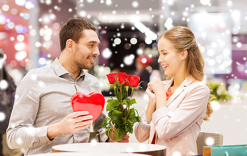 love, romance, valentines day, couple and people concept - happy young man with red flowers giving present to smiling woman at cafe in mall with snow effect