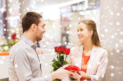 love, romance, valentines day, couple and people concept - happy young man giving red roses and present to smiling woman at cafe in mall with snow effect