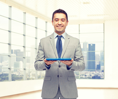 business, people and technology concept - happy smiling businessman in suit holding tablet pc computer over office room background