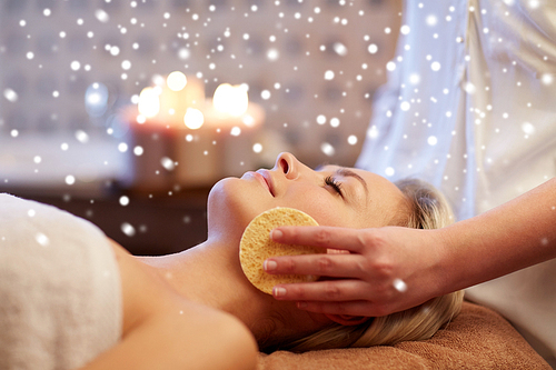 people, beauty, spa, skin care and relaxation concept - close up of beautiful young woman lying with closed eyes and having face massage with sponge in spa with snow effect