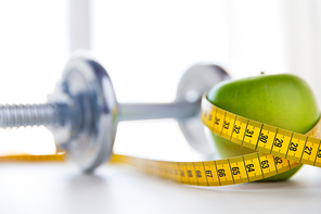 sport, fitness, diet and objects concept - close up of dumbbell and green apple with measuring tape