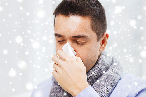 healthcare, flu, people, rhinitis and medicine concept - close up of ill man blowing his nose with paper napkin at home over snow effect