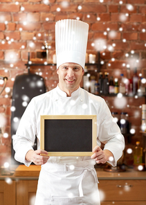 cooking and people concept - happy male chef cook with black blank menu chalk board in kitchen in restaurant kitchen over snow effect