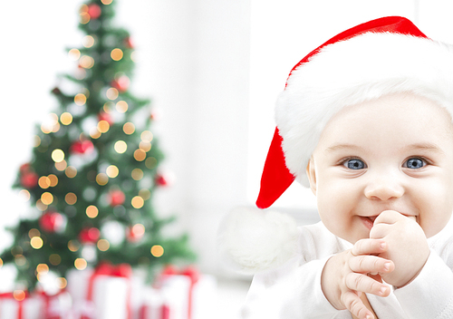 holidays, babyhood, childhood and people concept - happy baby in santa hat over christmas tree lights and gifts background