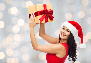 people, holidays, christmas and celebration concept - beautiful sexy woman in red dress and santa hat with gift box over holidays lights background