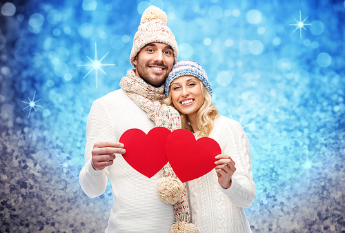 love, valentines day, couple, christmas and people concept - smiling man and woman in winter hats and scarf holding red paper heart shapes over blue glitter and holidays lights background