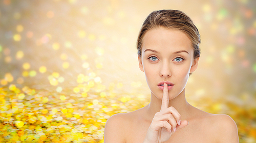people, silence, secret, gesture and beauty concept - beautiful young woman holding finger on lips over golden glitter or holidays lights background