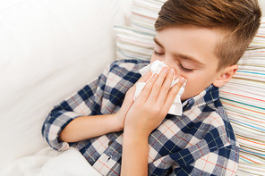 childhood, healthcare, rhinitis, people and medicine concept - ill boy with flu lying in bed and blowing his nose at home