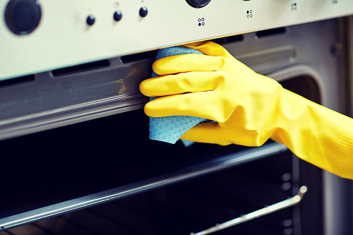 people, housework and housekeeping concept - close up of woman hand in protective glove with rag cleaning oven at home kitchen