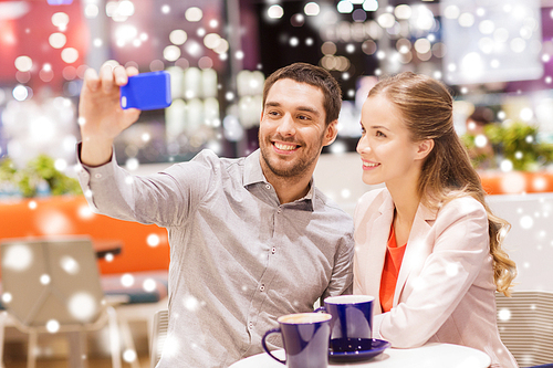 sale, shopping, consumerism, technology and people concept - happy young couple with smartphone taking selfie and drinking coffee or tea at cafe in mall with snow effect