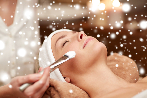 people, beauty, spa, cosmetology and skincare concept - close up of beautiful young woman lying with closed eyes and beautician hand applying facial mask by brush in spa salon with snow effect