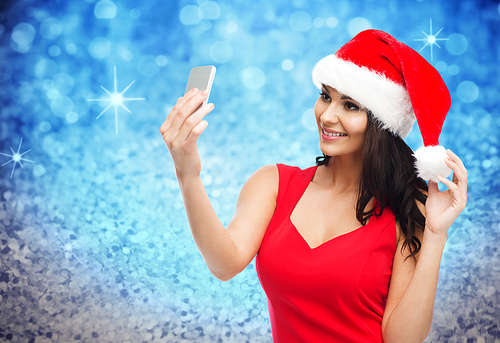 people, holidays, christmas and technology concept - beautiful  woman in red santa hat taking selfie picture by smartphone over blue glitter and lights background